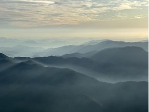 RAIOS DE SOL E NÉVOA SOBRE SILHUETA DE MONTANHAS DE UM PICO
